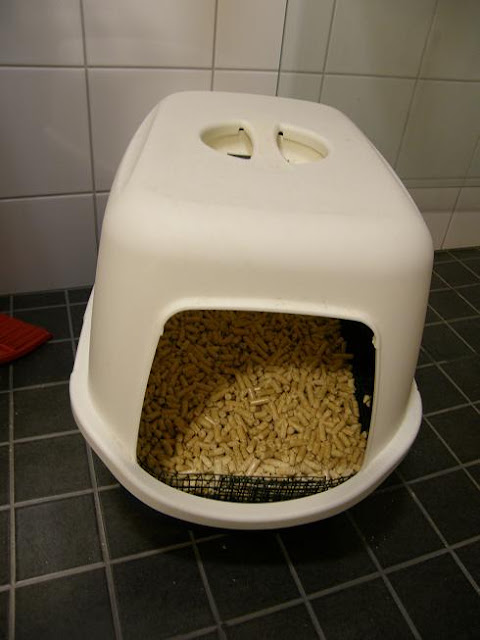 White covered litter box filled with tan pellet litter on dark tiled floor